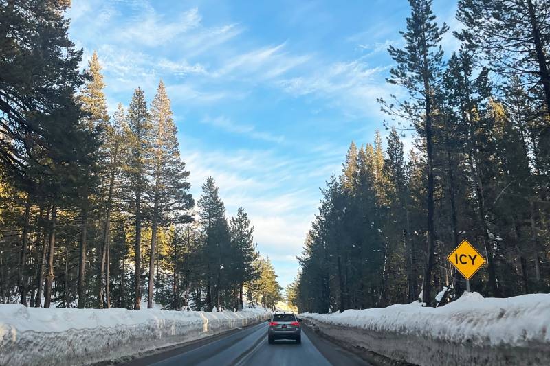 A photo of a road taken from inside a car, with banks of snow on either side, one car up ahead and a bright blue sky and pine trees.