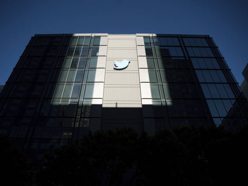 An image of a glass paneled office building with a blue outline of a bird on the side. The building is covered in shadows except for a square of light that falls on the blue bird.