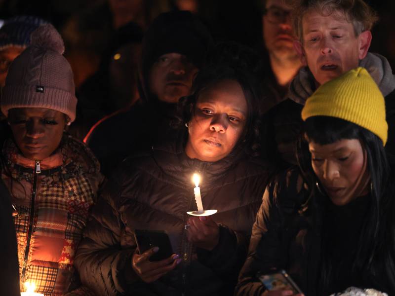 A woman with darker skin is holding a candle near her face. Around her are a group of people. They all appear to be standing outside in the dark, and everyone is wearing warm clothing.