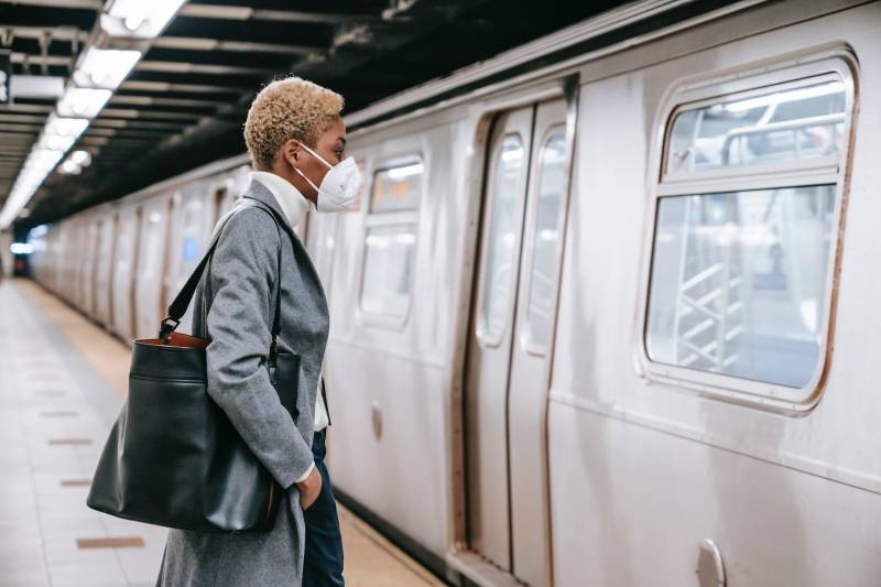 A person with dark skin and blonde hair wearing a white N95 mask and a sharp grey business-style overcoat waits on a subway platform as a train approaches