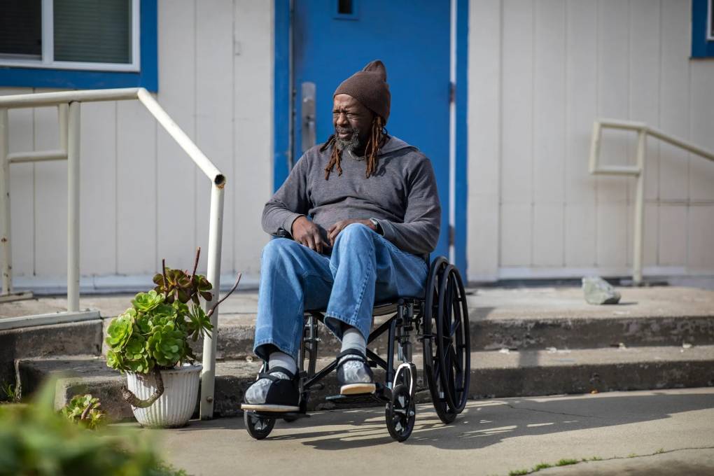 An older Black man in a wheelchair outsidea white house with a blue window and door.