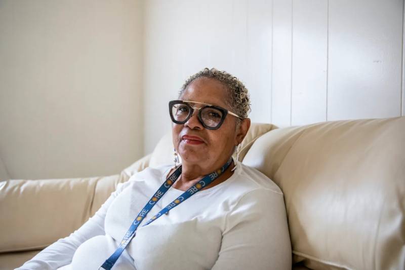 An older Black woman with graying hair and a white shirt and glasses looks at the camera while seated on a white couch.