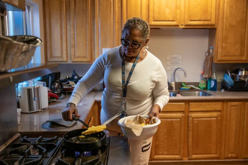An older Black woman cooks food in a trailer kitchen.