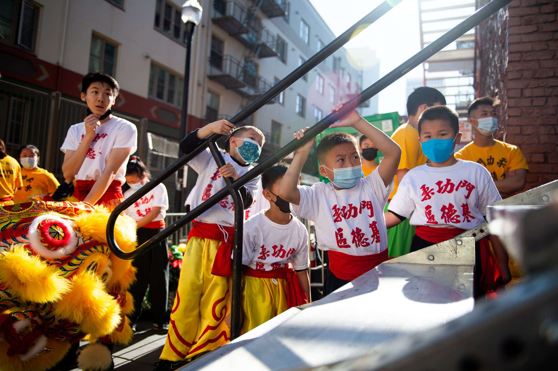 young children wearing white Yau Kung Moon shirts and gold leggings with red sashes hold onto a railing while looking toward an outdoor stage