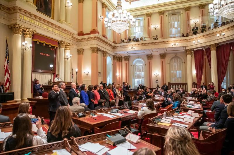 A line of people in the California State Capitol Senate chambers stand in front of a room full of other people who are seated.