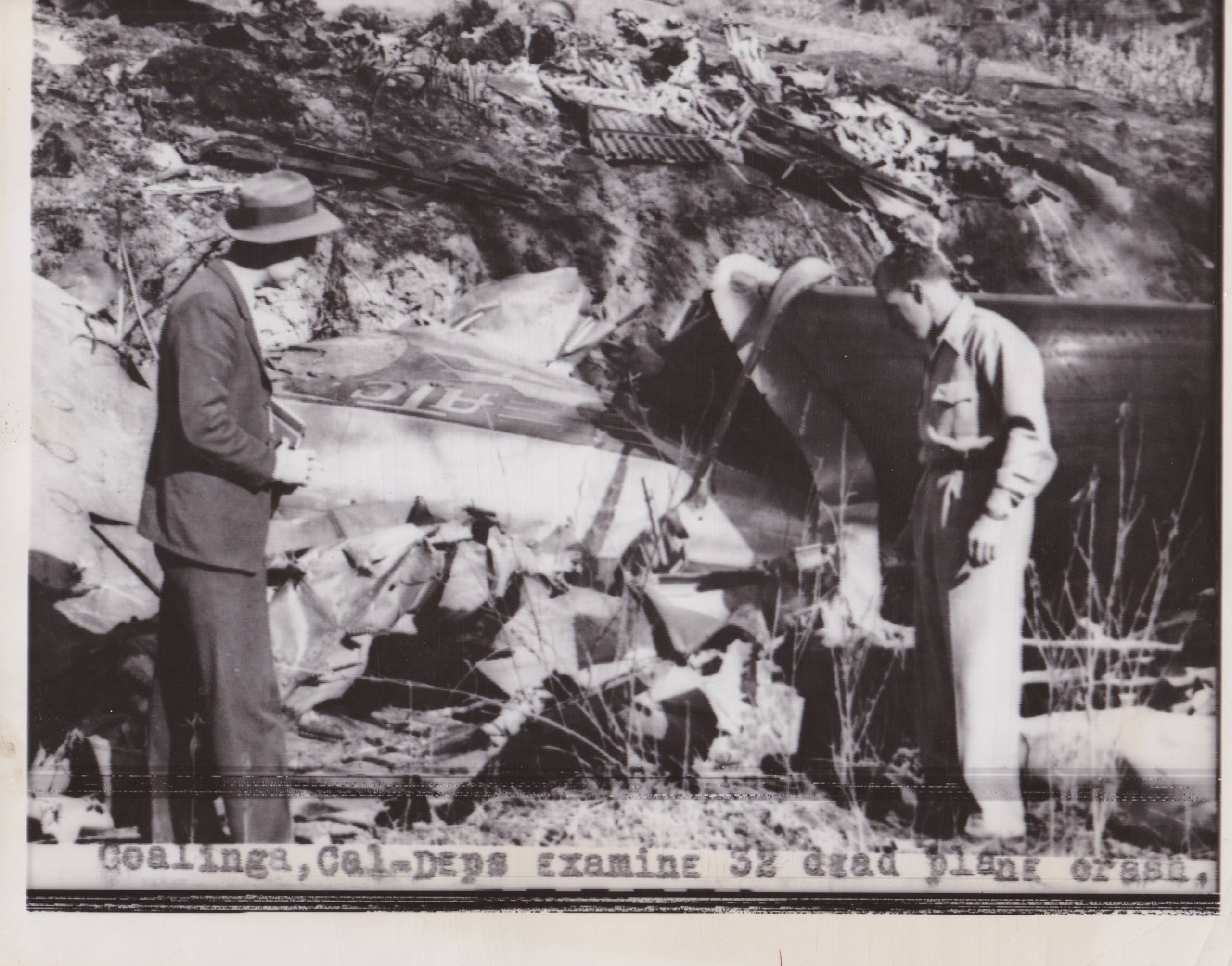 Two men stand in front of a plane wreckage site.