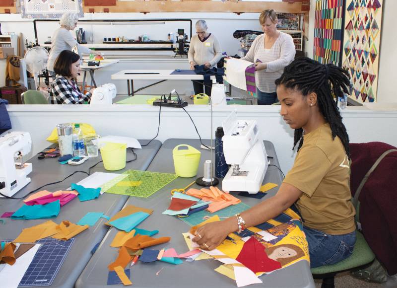 A Black woman works on a quilt at a table in the foreground with four white women working on quilts behind her in a workshop.