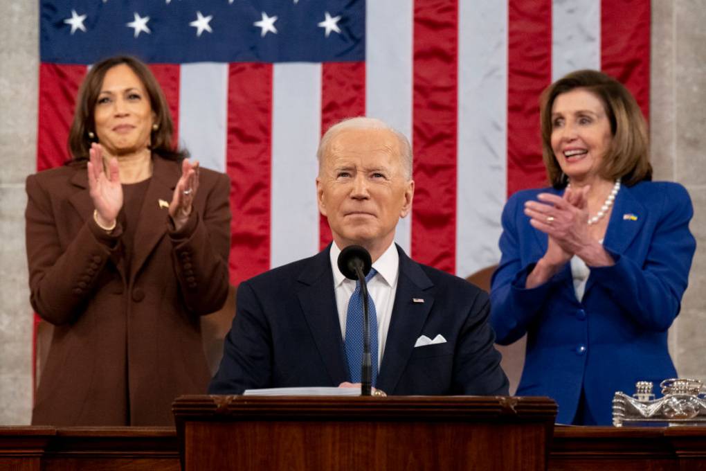 A man dressed in a business suit is behind a podium with a woman dressed in a brown suit to his left and a woman dressed in a blue suit on his right who are both clapping. They are all behind a large American flag.