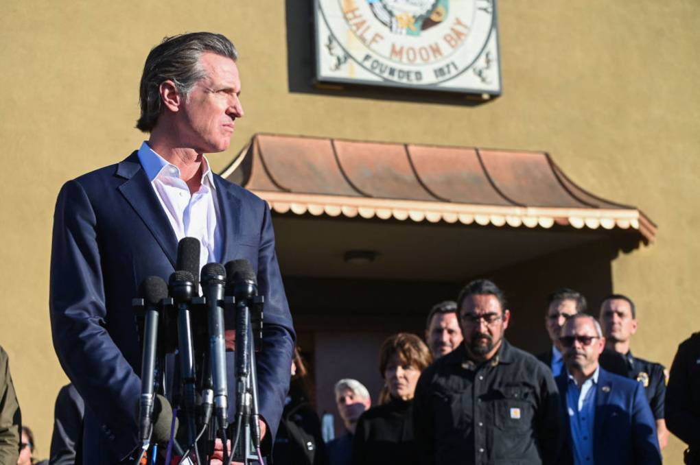 Gov. Gavin Newsom addresses a crowd in front of a building with a 'Half Moon Bay' sign on the facade.
