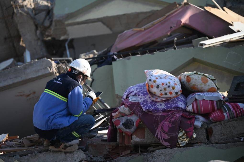 A rescue worker with a white helmet crouches beside rubble as he looks at his phone.
