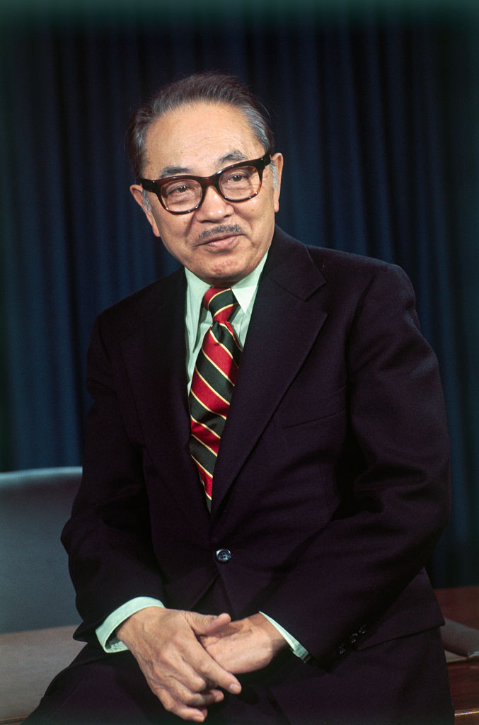 A middle-aged Asian man, with a slight, pleasant smile, poses sitting against the front of a desk or table He has dark gray hair combed neatly back, a thin gray moustache, and dark brown thick-framed glasses. He wears a dark blue suit, a powder green collared shirt, and a bright striped red and olive tie, with smaller yellow stripes. His hands are clasped lightly below his suit button.