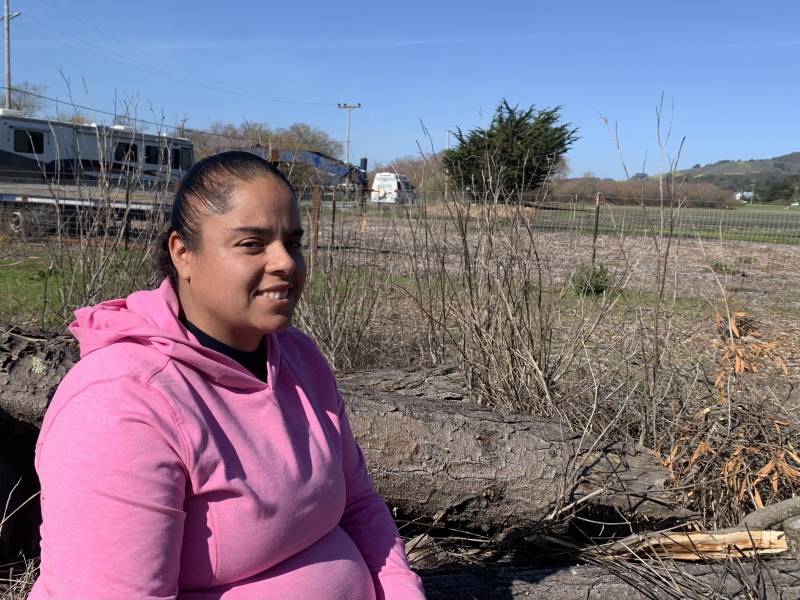 A Latina woman with a pink hoodie smiles at the camera while working on a farm.