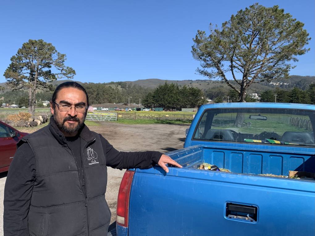 A Latino man with beard and glasses rests his hand on the back of a blue pickup truck as he smiles to the camera with trees and hills in the background.
