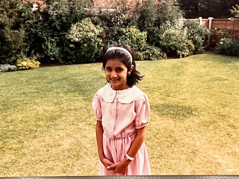 A young Pakistani-American girls stands on the grass smiling at the camera. She is wearing a pink dress with a white collar, and her hair pulled back with a headband. 
