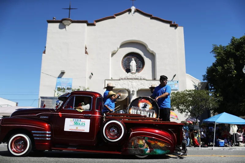 A dark red lowrider truck is almost center frame, with a group of percussionists standing in the truck's bed. In the background, a church.
