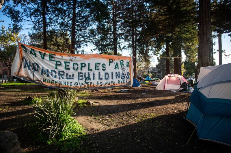 A spraypainted sign reads "Save Peoples Park, No More Buildings" near tents in a park.