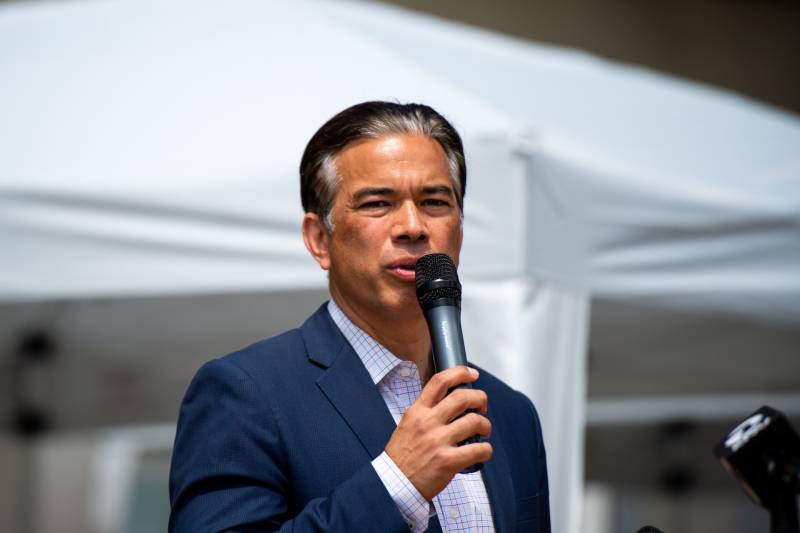 Rob Bonta stands center frame wearing a navy blue suit and white dress shirt while speaking into a microphone. Behind him is a large white canopy.