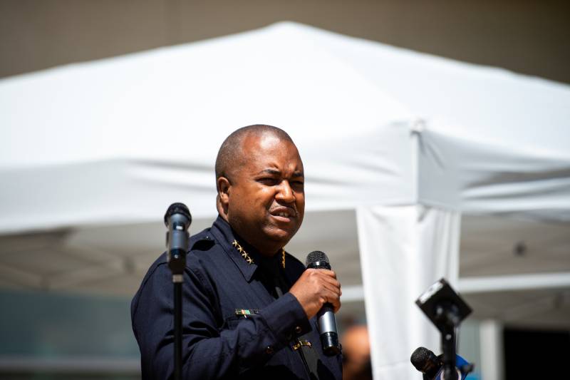 A bald Black man in a blue police uniform stands outside in the sun holding a microphone and squinting