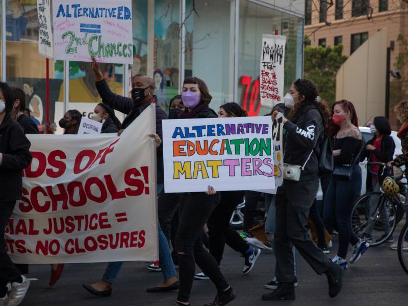 A group of protesters hold signs against school closures as they march on a city street.