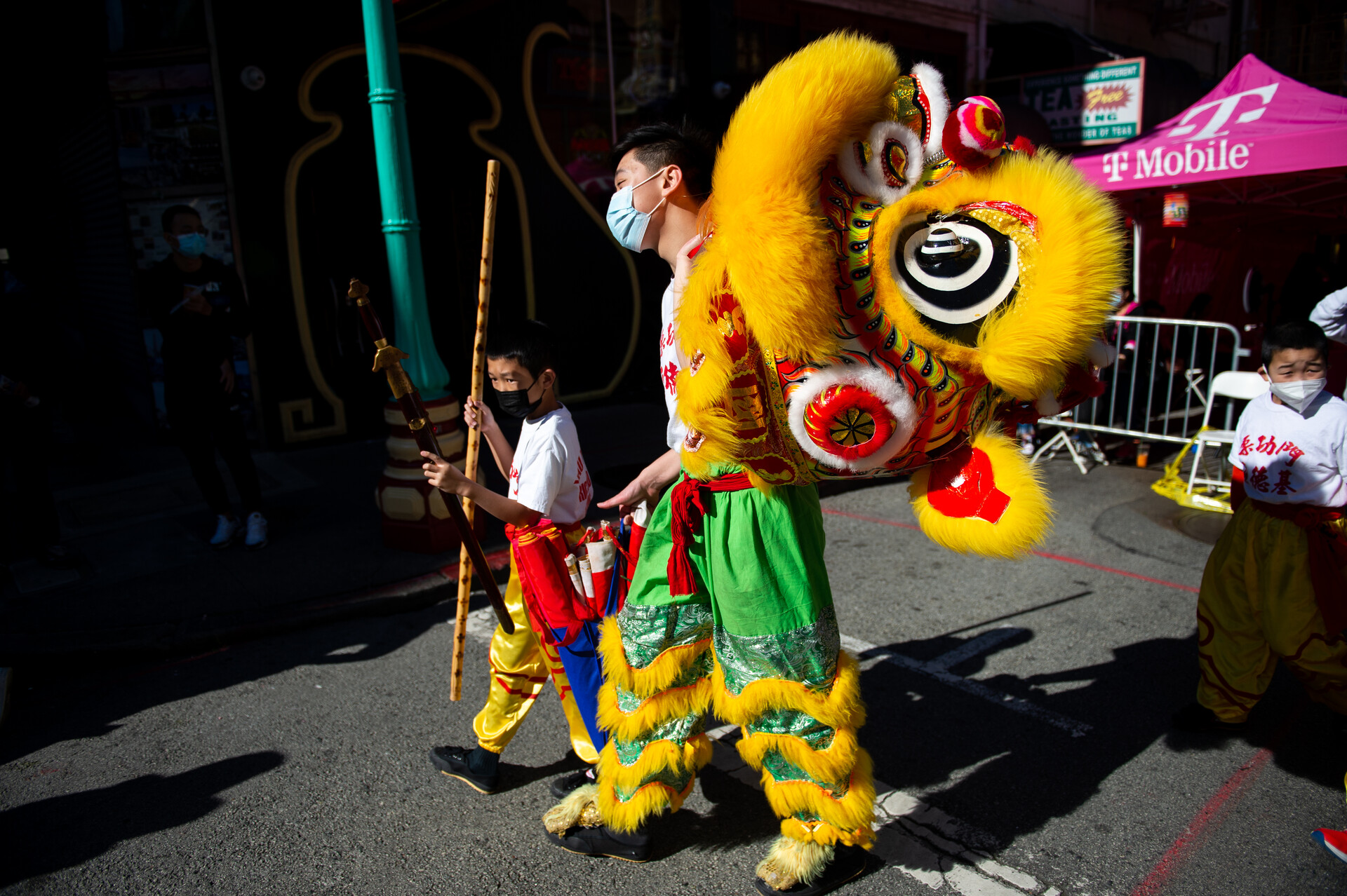 An Asian man carrying a large yellow dragon head, wearing green leggings with gold trim, walks with a young boy carrying a staff outside in Chinatown