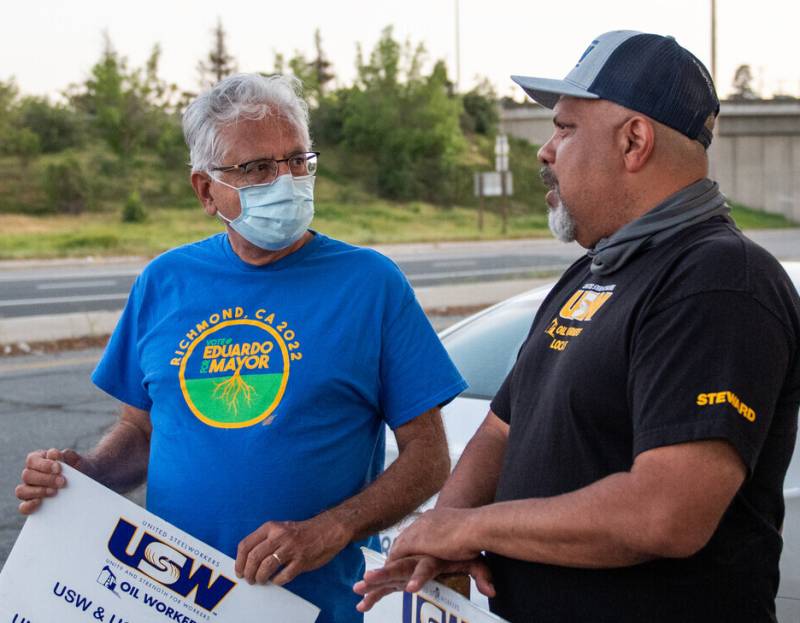 An older Latino man in a blue shirt holds a strike placard as he talks to a striker, a man in a black shirt with a cap on.