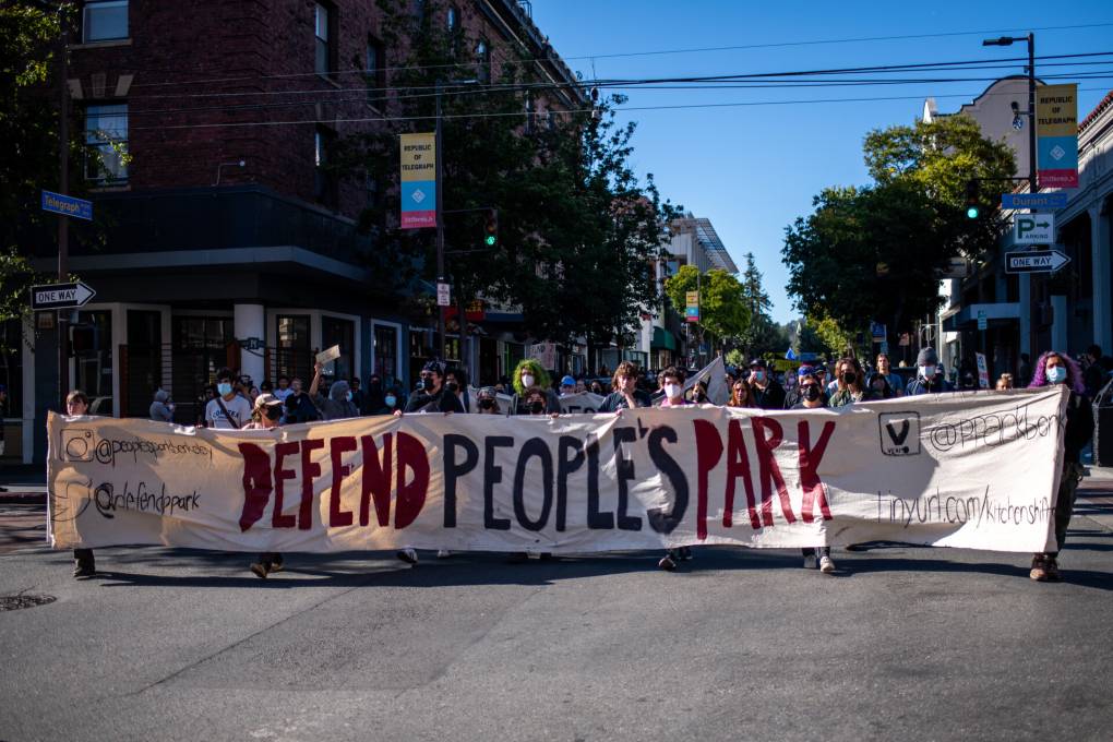 A crowd marches toward the camera, with those in front holding a large banner reading "Defend People's Park"