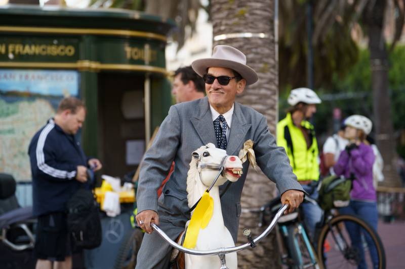 A man with a wide-brimmed hat, sunglasses, and a suit sits astride his custom bicycle constructed of a playground-style spring horse mounted to a BMX bike.