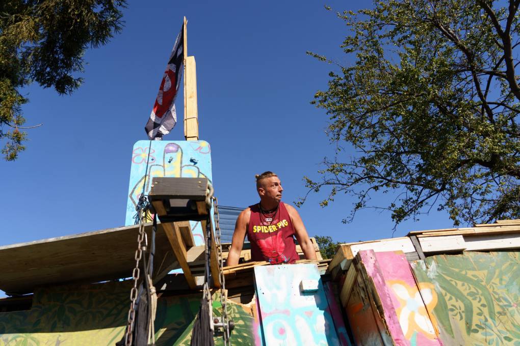 A man wearing a sleeveless red t-shirt looks out from on top of a makeshift structure.