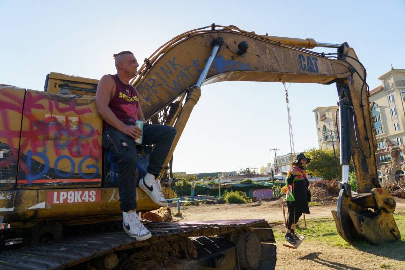 A man wearing a sleeveless T-shirt sits on the side of an excavator as a person stands on a makeshift swing hanging from the arm of the construction vehicle.
