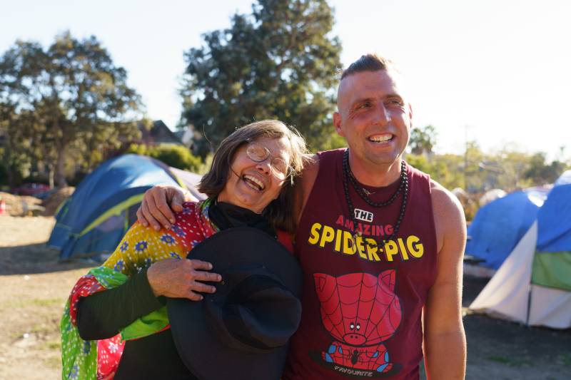 A man wearing a red sleeveless shirt that says "The Amazing Spider-Pig" with an illustration puts his arm around a person wearing glasses and dark clothes with a light-colored blanket over their shoulder.