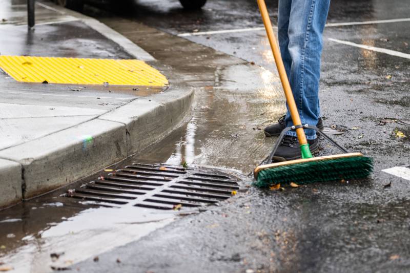 Close up image of a storm drain on a street corner. Someone is sweeping debris away from it.