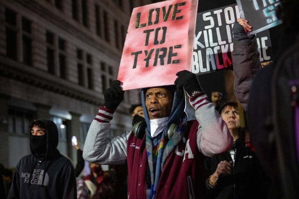 A Black man wearing a white and maroon jacket and a hoodie holds a hand-made sign reading 'Love to Tyre' above his head, with another sign reading 'Stop Killing Black People' visible in the back, with the buildings of downtown in the background