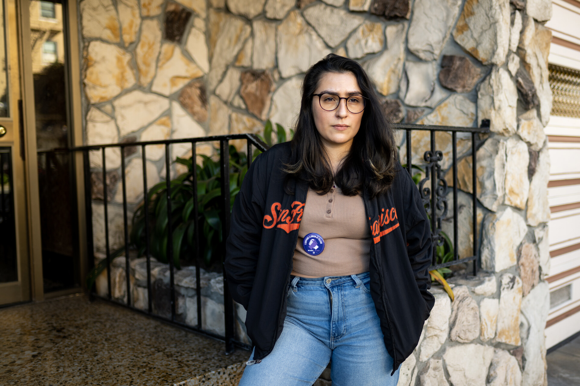 A woman with glasses and a SF Giants jacket stands in front of stone building.