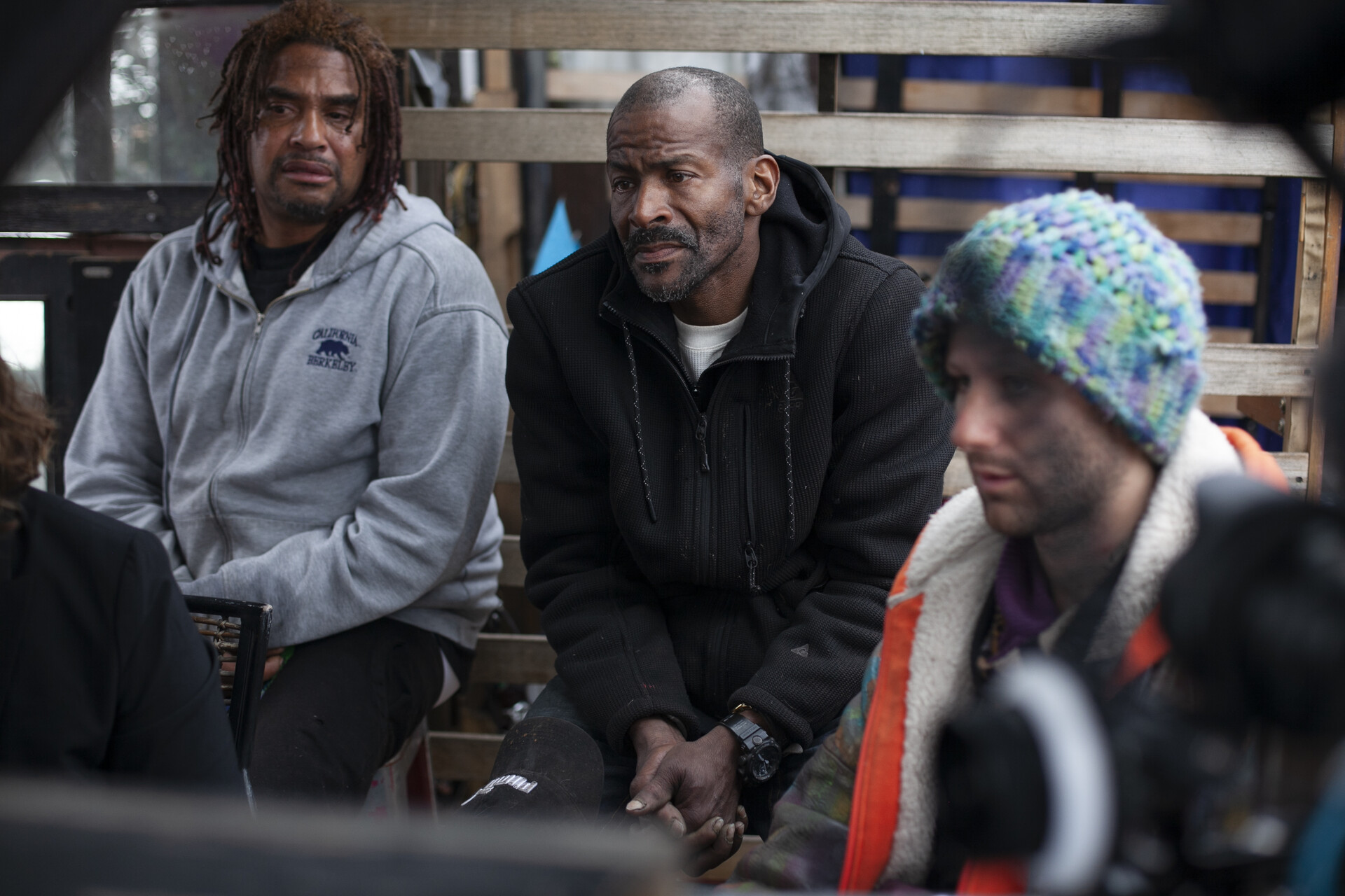 Three people with concerned expressions sit together listening: a Black man with dreads wearing a gray hoodie sits next to a Black man wearing a black hoodie, and a white man wearing a multicolored beanie