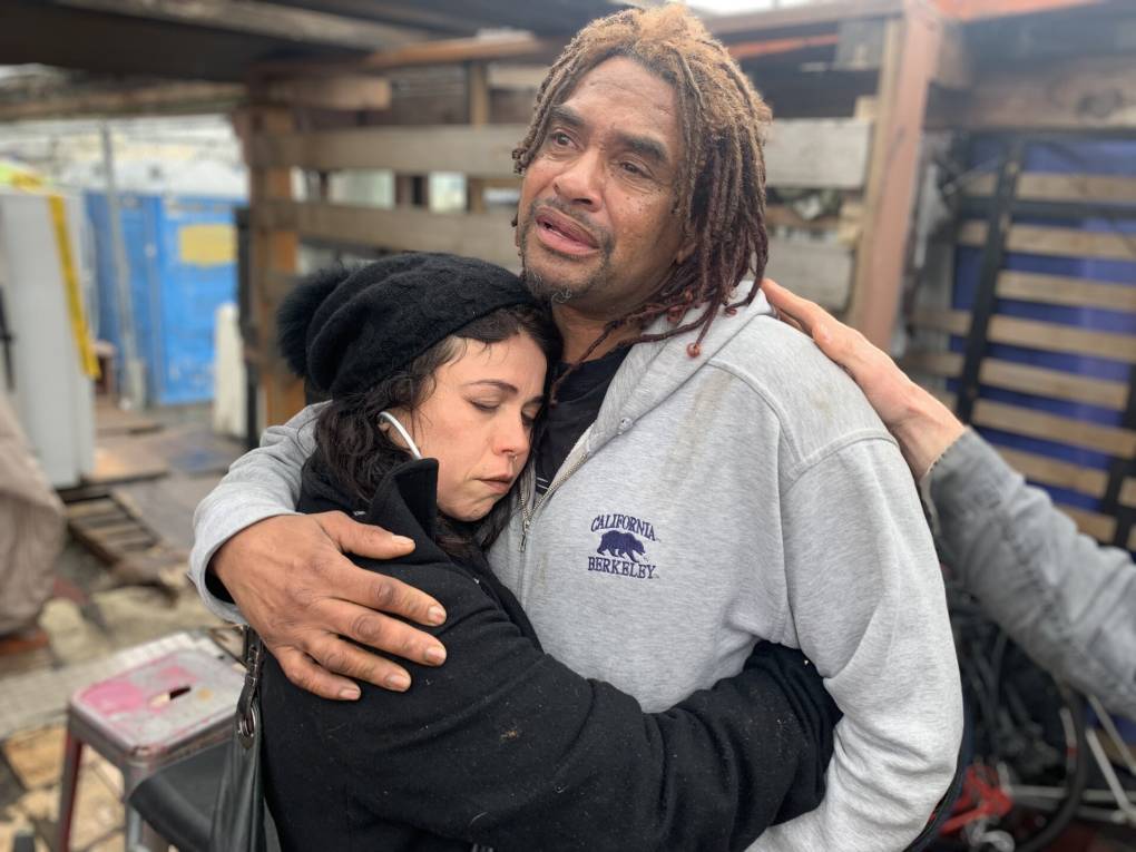 A white woman with her eyes closed wearing a black sweatshirt and beanie hugs a tearful Black man wearing a gray sweatshirt