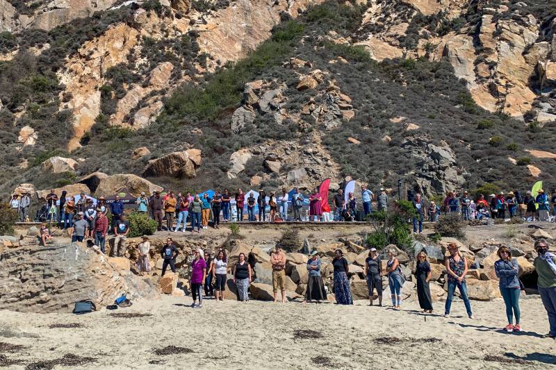 Hundreds of people form a line at the base of a large rock outside.