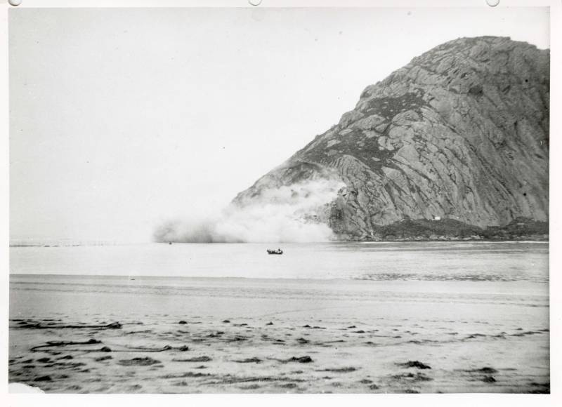 An archival black and white photo of a section of a large rock being blown up.