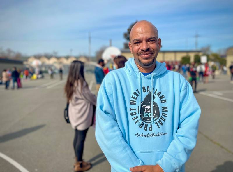 A Black man with clean shaven head and wearing a blue hoodie that reads "Protect West Oakland Mural" smiles at the camera with people in the background.