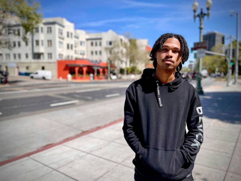 A young Black man with a black hoodie and shoulder-length black twists looks at the camera while standing on a sidewalk in an urban street.
