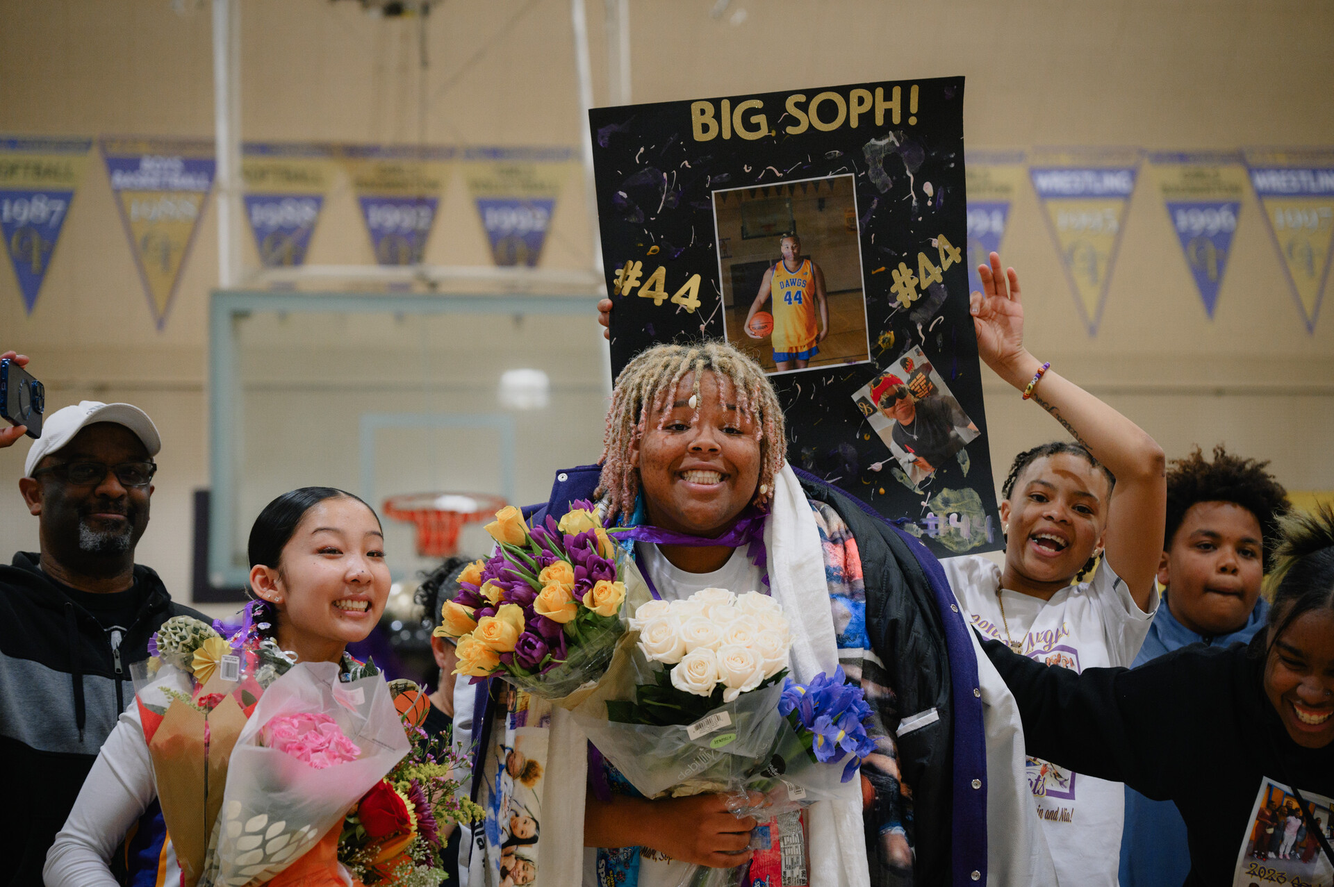 Two broadly smiling teenage girls, one shorter, Asian, with dark hair, one taller, Black, with blonde braids, stand in the middle of a basketball court holding flowers surrounded by friends and family. Children behind the girl in the center hold a homemade sign reading 'Big Soph!'