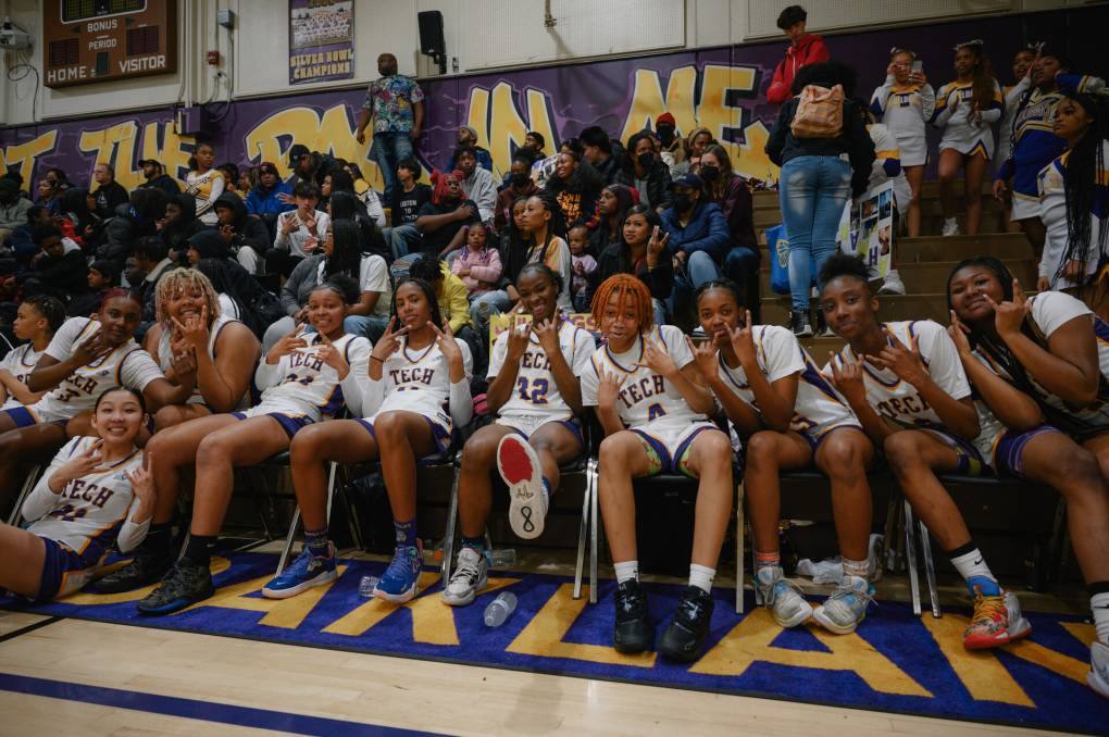 Photo of a row of smiling, mostly Black teenage basketball players wearing white jerseys courtside, smiling and signaling with their hands