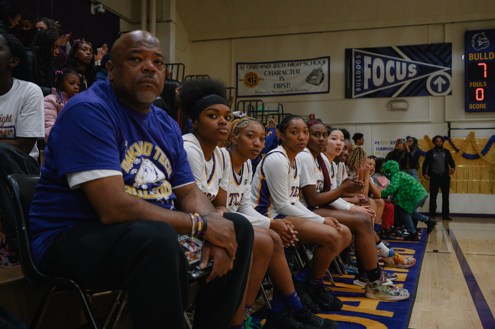 Perspective courtside photo of a coach wearing a purple shirt sitting next to six or seven high school girl basketball players wearing white jerseys