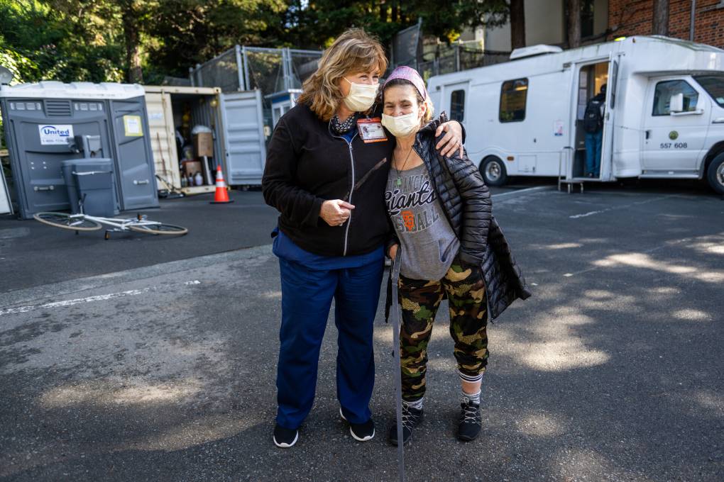 Two women with light-coilored skin and wearing face masks stand in front of a trailer, with their arms around each other.