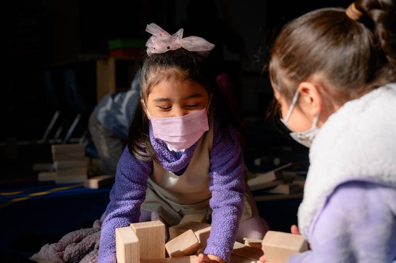 Two girls play with blocks on a carpet in a classroom.