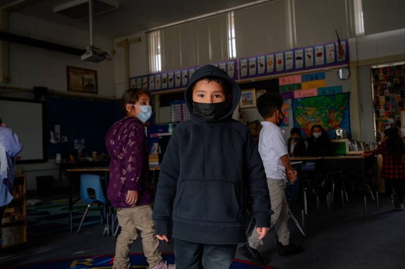A young boy with a face mask looks at the camera in a classroom with other kids playing behind him.