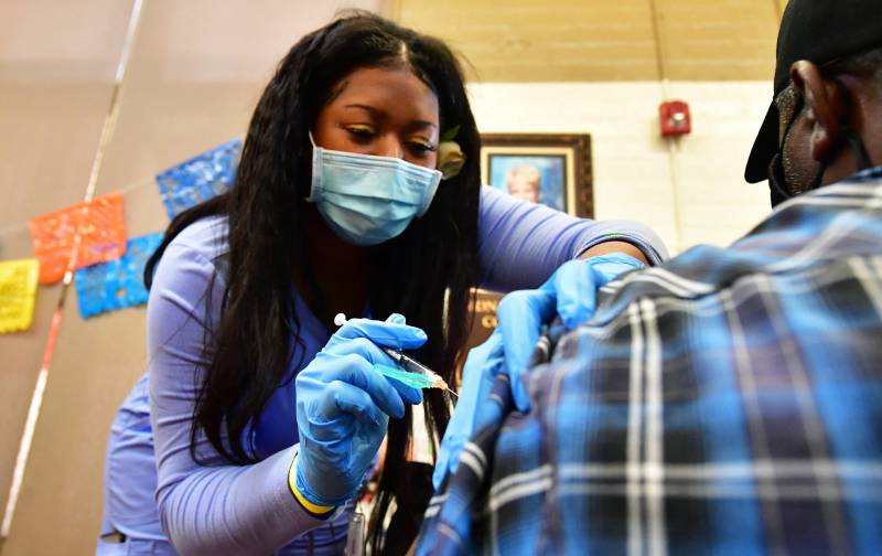 A Black woman wearing a blue surgical mask and a blue shirt faces the camera, leaning over to administer a vaccine to a person wearing a blue checked shirt, whose back is to the camera.