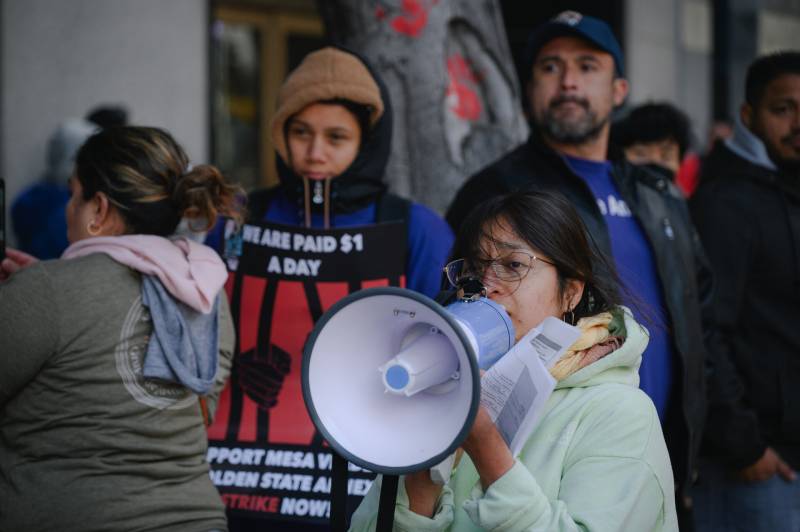 A Latina woman speaks into a megaphone with male and female protesters behind her.