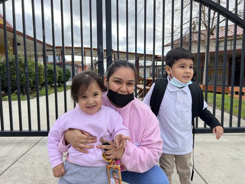 A Latina woman in a pink sweater poses with her two young children outside of a school gate.