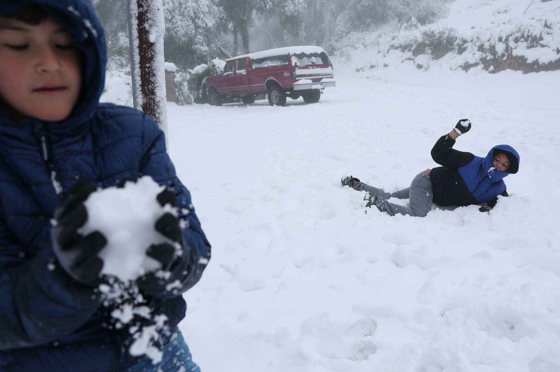 two children wearing blue coats have a snowball fight - one in the foreground packs a snowball facing the camera as one in the background lies horizontal in the snow, which appears to be a foot deep