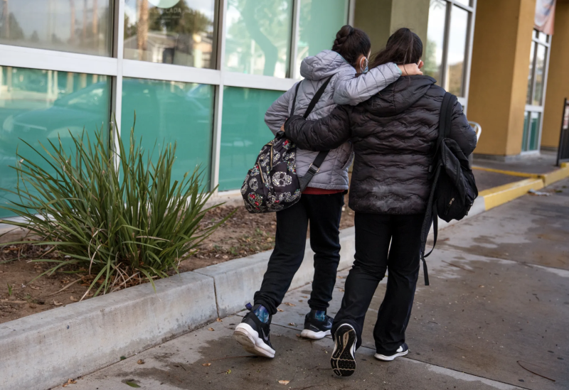 Two young people wearing gray and black jackets are walking on a sidewalk down the street with their arms around each other's shoulder and back.